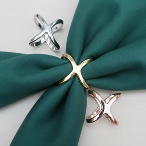 May include: An X-shaped napkin ring in gold, silver, and rose gold. The gold ring is holding a dark teal napkin. Two other rings are placed on a white surface. These decorative rings are perfect for a formal dinner setting.