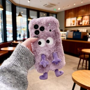 Op de afbeelding: Een pluizig, lavendelkleurig telefoonhoesje met een monsterontwerp. De hoes heeft grote, wiebelende ogen, kleine paarse voetjes en een zachte, pluche textuur. De hoes is ontworpen voor een smartphone en heeft uitsparingen voor de camera en andere functies.