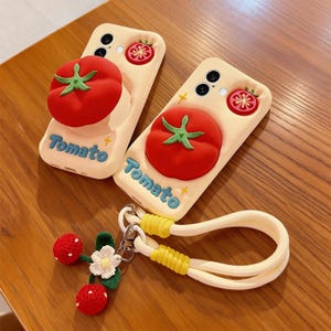 May include: Two cream-coloured phone cases featuring a red tomato design with green stems. The word "Tomato" is written in blue. A matching keychain with red cherry-like beads and a white flower is also present. The cases are on a wooden surface.