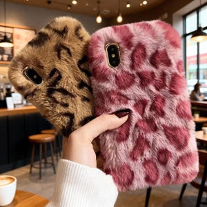 Puede incluir: Dos fundas de teléfono peludas. Una es marrón con manchas marrones oscuras, la otra es rosa con manchas rosas más oscuras. Ambas fundas tienen un marco dorado para la lente de la cámara.