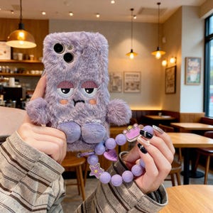 Puede incluir: Una funda de teléfono peluda, de color lavanda, con una cara de monstruo bordada, con ojos somnolientos y un ceño fruncido. La funda tiene pies adjuntos y una correa de muñeca de cuentas a juego con un dije de corona y detalles de estrella.