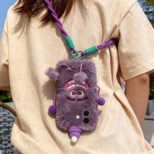 May include: A purple, fluffy phone case with a monster face design, featuring large eyes, a wide grin, and small pom-poms. The case has a matching purple and green strap. The phone case is designed to be worn around the neck.