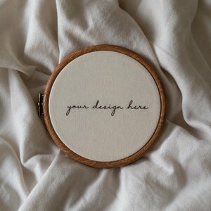 May include: A circular wooden embroidery hoop containing a piece of off-white fabric stretched taut. The fabric features the black embroidered text "your design here" in a cursive script centered within the frame of the hoop.