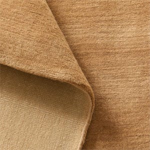 May include: A close-up of a tan-colored rug with a rolled-up corner, revealing the underside. The rug has a textured surface and a neutral color palette, suitable for various interior design styles.