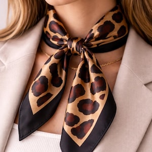 Puede incluir: Un pañuelo de seda con estampado de leopardo. El pañuelo presenta un fondo beige con manchas marrones y bordes negros. Está anudado alrededor del cuello, creando un accesorio elegante. El pañuelo se combina con una blazer y un collar dorado.