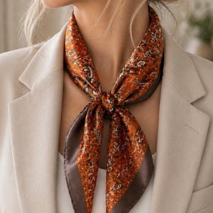 Op de afbeelding: Een oranje paisley zijden sjaal met een donkerbruine rand is om de hals geknoopt. De sjaal wordt gedragen over een witte top en een beige blazer. Het patroon van de sjaal bevat witte en donkerbruine details.