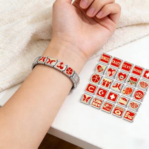 Puede incluir: Una pulsera de dijes de acero inoxidable con dijes rojos y dorados. Se muestran dijes adicionales con varios diseños y palabras como "Bingo", "Crazy" y "Sexy" junto a la pulsera.