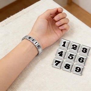 Puede incluir: Una pulsera plateada con los números negros 0-3 se lleva en la muñeca. Se muestran nueve dijes plateados adicionales con los números 1-9. Los números están en una fuente negrita sin serifa. El fondo es de tela beige claro.