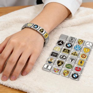 May include: A stainless steel charm bracelet with various car brand logos, including BMW, Porsche, and Audi. The bracelet is on a wrist, with additional charms displayed nearby. The charms are rectangular with colorful enamel designs.