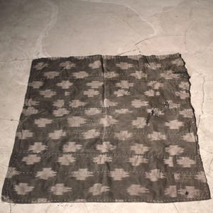 May include: A dark gray textile with a repeating pattern of light gray geometric shapes. The edges are stitched with white thread. The textile is rectangular and appears to be handmade.