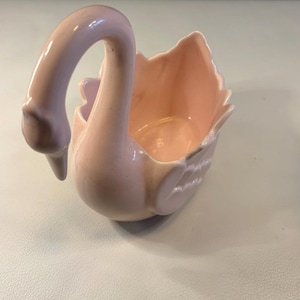 Könnte beinhalten: Ein hellrosa Keramik-Schwan-Pflanzgefäß. Der Schwan hat einen langen, gebogenen Hals und ein detailliertes Flügeldesign. Das Innere ist passend rosa, wodurch ein zusammenhängendes und dekoratives Stück entsteht. Ideal für kleine Pflanzen oder als dekoratives Element.