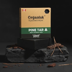 Può includere: Una scatola di sapone naturale Organisk Pine Tar con una bandiera canadese. La scatola è beige con dettagli verdi e neri. Una saponetta nera è appoggiata su una pietra grigio scuro. Il sapone è Oatmeal & Sand Heavy Grit, peso netto 125 g.