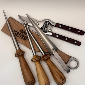 Puede incluir: Un conjunto de herramientas de jardinería, que incluye tres punzones con mango de madera, unas tijeras de podar y una herramienta cónica. Las herramientas son de metal y madera, con los punzones y las tijeras con un acabado plateado. Las tijeras tienen mangos de color marrón oscuro. Se ve una funda de madera.