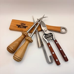 May include: A collection of gardening tools, including a pair of pruning shears with wooden handles, two awls, and a wooden handle with a hole. The tools are arranged on a white surface. The wooden handle has the text "NORTHWEST WILLOW".