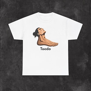 May include: A white short-sleeve t-shirt featuring a graphic illustration of a human head and neck transitioning into a foot, with tears streaming down the face. The word Toedo is printed in black text below the graphic illustration.