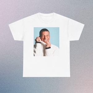 Puede incluir: Una camiseta blanca de manga corta con un estampado gráfico rectangular centrado. La imagen muestra a una persona sonriente con un suéter blanco de punto trenzado con ribetes negros en los puños, sobre un fondo azul claro.