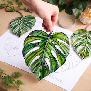 Peut inclure: Ornements faits à la main en forme de feuilles de Monstera vertes et blanches avec un motif de veines détaillé. Les feuilles sont présentées sur un papier blanc avec un contour de feuille. Les feuilles sont attachées à une ficelle brune.