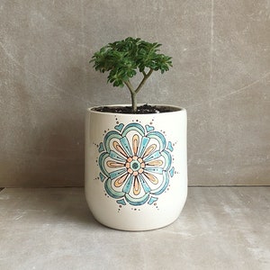 Puede incluir: Una maceta de cerámica blanca con un diseño de mandala azul, verde y naranja. Una pequeña planta suculenta verde está creciendo en la maceta.