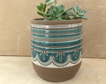 Könnte beinhalten: Ein brauner Keramiktopf mit einem gestreiften Design in Teal und Weiß und einem gewellten Rand in Teal und Weiß. Der Topf ist mit verschiedenen Sukkulenten gefüllt.