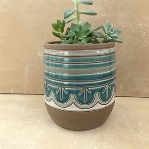 Puede incluir: Una maceta de cerámica marrón con un diseño a rayas azul verdoso y blanco y un borde festoneado azul verdoso y blanco. La maceta está llena de diferentes suculentas.