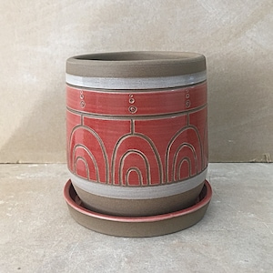 Può includere: Un vaso di terracotta rosso con un bordo bianco e un design geometrico. Il vaso ha un sottovaso abbinato.