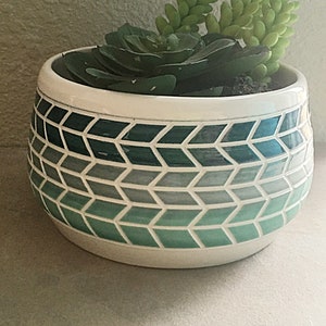 May include: A white ceramic planter with a geometric pattern in shades of green and blue. The planter is filled with a succulent plant.