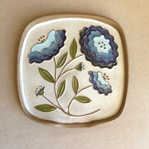 May include: A square ceramic plate with rounded corners featuring a hand-painted floral design. The plate has a cream-colored center and a textured brown rim. Three stylized flowers in shades of blue and light blue with green leaves.