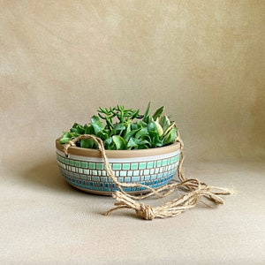 May include: A hanging planter made of ceramic with a blue and green mosaic design. The planter is filled with green succulents and has a natural twine rope for hanging.