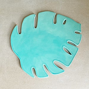 May include: A turquoise ceramic plate shaped like a leaf. The plate has a distinctive design with cut-outs around the edges, reminiscent of a tropical leaf.