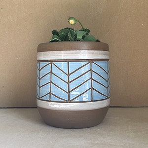May include: A light blue ceramic planter with a chevron pattern and a white rim. The planter is filled with a small green succulent plant.