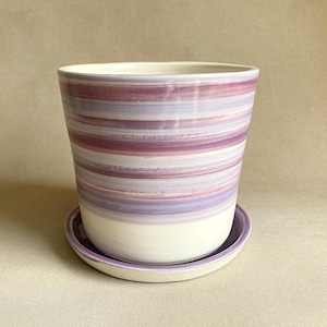 May include: A ceramic planter with a matching saucer. The planter is cylindrical with a wide opening and features horizontal stripes in shades of purple and white. The saucer is a matching purple.