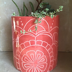 May include: A coral-colored ceramic planter with a white floral design. The planter is filled with green succulents.