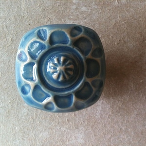 May include: A square ceramic knob with a blue glaze and a white floral design. The knob has a raised centre with a starburst pattern.