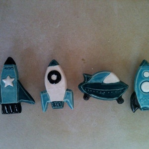 May include: Four ceramic space-themed figurines. There are two rockets, a spaceship, and a flying saucer. The rockets are teal, white, and black. The spaceship and flying saucer are teal and white.