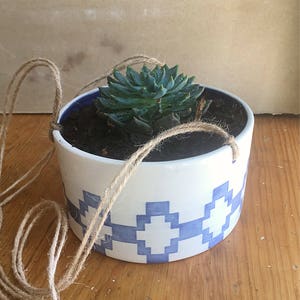 May include: A white ceramic hanging planter with a blue geometric pattern, featuring a small green succulent plant. The planter is suspended by a natural twine rope.