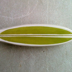 May include: A white ceramic surfboard with a green painted design and two white stripes.