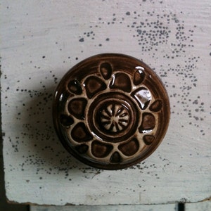 Peut inclure: Un bouton en céramique brun avec un motif floral blanc. Le bouton a un centre surélevé avec un petit cercle au milieu.