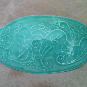 Può includere: Un piatto in ceramica color turchese con un motivo paisley bianco. Il piatto è di forma ovale e ha una finitura lucida.