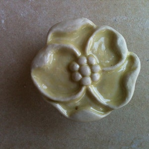 May include: A ceramic flower-shaped knob in a pale yellow color with a raised center detail. The knob has a smooth, glossy finish.