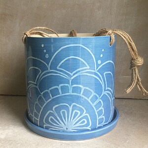 May include: A blue ceramic hanging planter with a white floral design and a matching saucer. The planter has a rope handle.