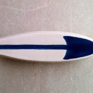 Puede incluir: Plato de cerámica blanco con forma de tabla de surf con una franja azul y un diseño de punta de flecha azul.
