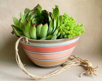 May include: A hanging planter made of ceramic with a striped design in shades of pink, orange, and white. The planter is filled with green succulents and has a brown twine rope for hanging.