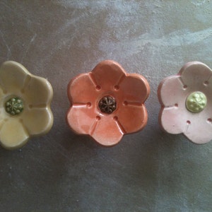 Peut inclure: Trois boutons en céramique en forme de fleur de couleur jaune, orange et rose. Chaque bouton a un petit centre vert en relief.