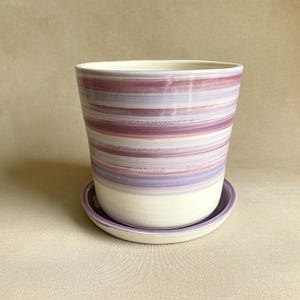 May include: A ceramic planter with a matching saucer. The planter is cylindrical with a wide opening and features horizontal stripes in shades of purple and white. The saucer is a matching purple.