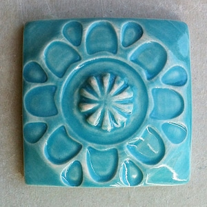 May include: A turquoise ceramic tile with a raised, embossed design of a flower with a central circle. The tile has a glossy finish.