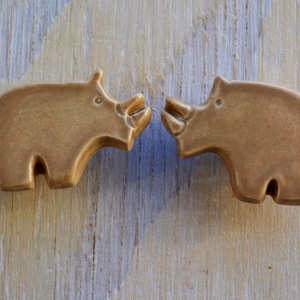 Może przedstawiać: Dwie brązowe ceramiczne figurki nosorożców, które patrzą na siebie. Figurki są proste i stylizowane, z gładkim, matowym wykończeniem.