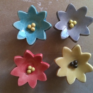May include: Four ceramic flower-shaped dishes with a central indentation. The dishes are in blue, purple, red, and yellow. Each dish has three small yellow or brown dots in the centre.