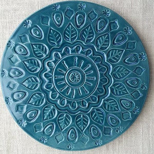 May include: A teal ceramic plate with a detailed, embossed design. The plate features a central sunburst pattern surrounded by leaves, eye-shaped elements, and floral accents. The plate is approximately 10 inches in diameter.