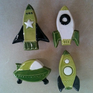 May include: Four ceramic rocket and spaceship ornaments in green, black and white. The rockets have black fins and white details. The spaceship is green with a white top and black wheels.