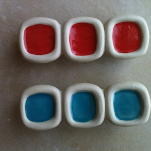 Puede incluir: Tres azulejos de cerámica blanca con bordes redondeados, cada uno con un cuadrado hundido. Los cuadrados están rellenos de esmalte rojo y azul.